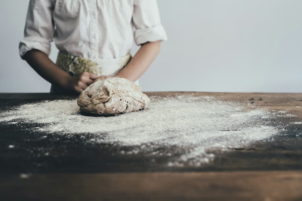 Een bakker in een wit overhemd aan een houten tafel, een bol deeg in een ruime laag bloem, in zacht ochtendlicht